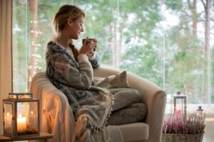 Winter, warm, Heat, Temperature, coffee, tea, blanket, Young beautiful woman sitting home in the chair by the window with cup of hot coffee wearing knitted warm sweater. Cozy room decorated with lanterns and candles.